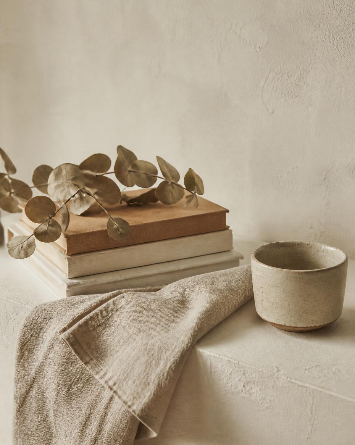 A stack of books, linen and a ceramic cup on a cream surface